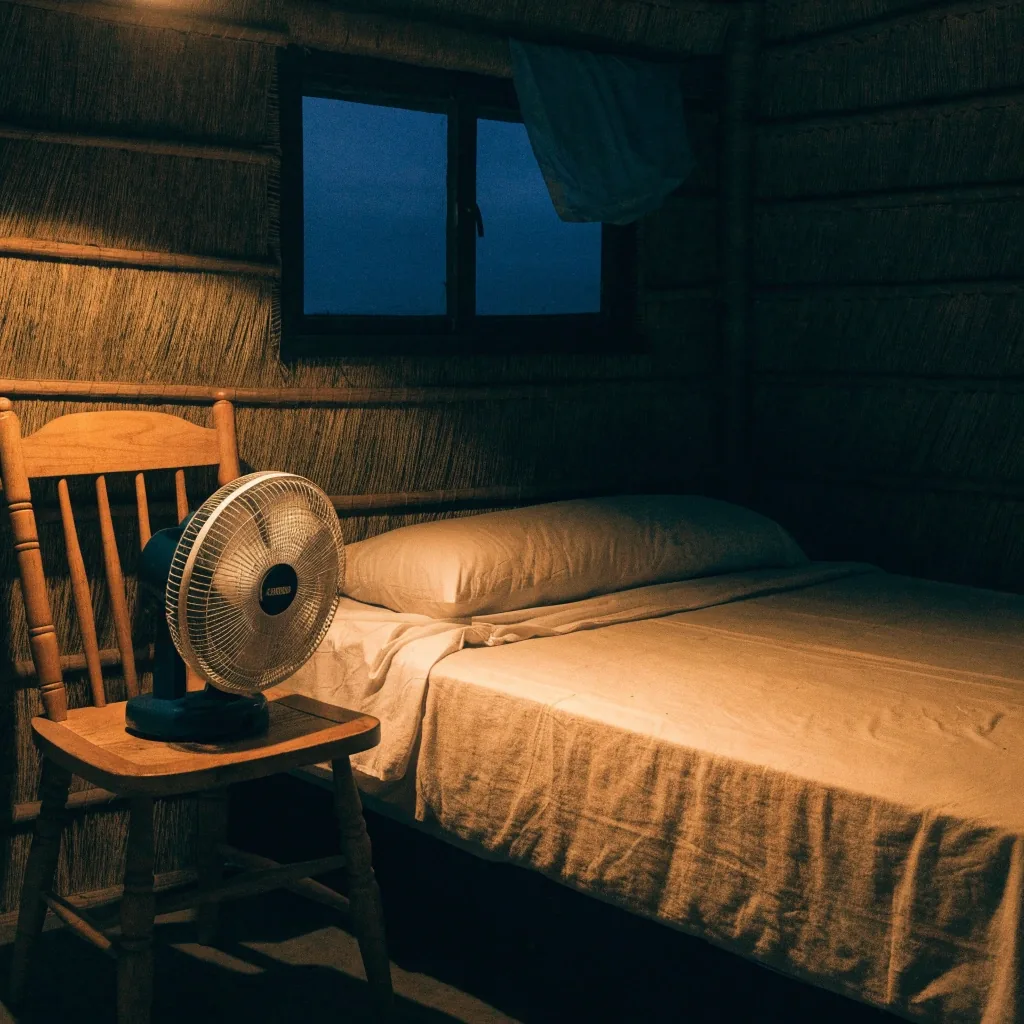 A simple shared bedroom with a fan beside a bed and a cloth over the window corner